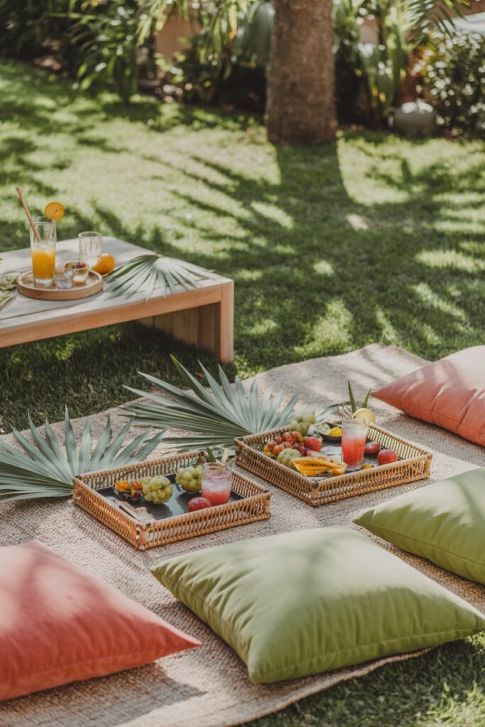 tropical garden picnic with palm leaf decorations, rattan trays filled with fruit, soft green and coral cushions layered on grass, wooden low table with tropical drinks, sunlight filtering through trees creating dappled shadows, airy outdoor atmosphere, cozy seating arrangement, warm natural lighting, relaxed vacation-inspired garden setting captured with vibrant yet soft tones.